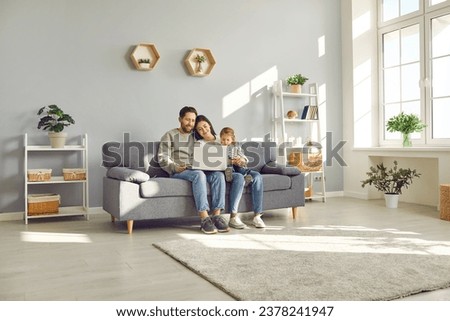 Full length photo of a happy young family with little child girl sitting on the sofa using modern laptop together. Parents resting on couch watching funny cartoon online or talking on video call.