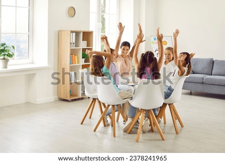 Happy children sitting in circle in classroom with friendly smiling school teacher or psychologist woman during group therapy. Kids girls raising hands up to ask or answer a question during meeting. Royalty-Free Stock Photo #2378241965