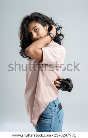A young girl stands against a white background, confidently holding a camera behind her and capturing photos, her poised silhouette capturing a sense of curiosity and creativity.