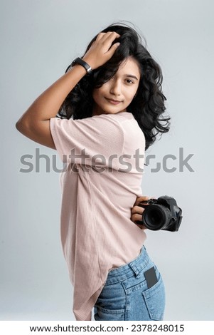 A young girl stands against a white background, confidently holding a camera behind her and capturing photos, her poised silhouette capturing a sense of curiosity and creativity.
