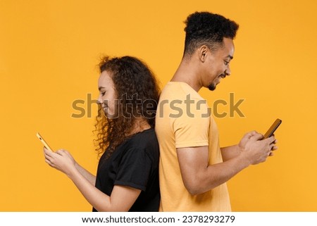 Side view young couple two friends together family african students woman man 20s in black t-shirt holding using mobile cell phone stand back to back isolated on yellow background studio portrait