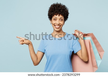 Young woman of African American ethnicity wearing casual clothes hold in hand paper package bags after shopping point aside isolated on plain light blue background. Black Friday sale buy day concept