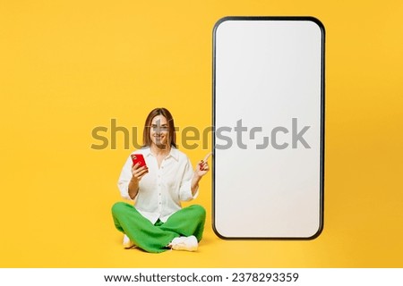Full body young smiling caucasian happy woman she wears white shirt casual clothes sit point on big huge blank screen mobile cell phone with area using smartphone isolated on plain yellow background