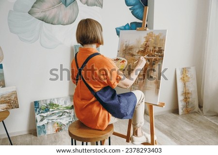 Full body back rear view elderly artist woman 50 years old wearing casual clothes sitting near easel with painting artwork paint spending free spare time in living room indoor. Leisure hobby concept