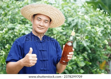 Asian man farmer wears hat, blue shirt and holds bottle of honey at his garden. Concept, Organic agriculture product from bee raising farm or nature sources. Thai local lifestyle. Natural beverage. Royalty-Free Stock Photo #2378299709