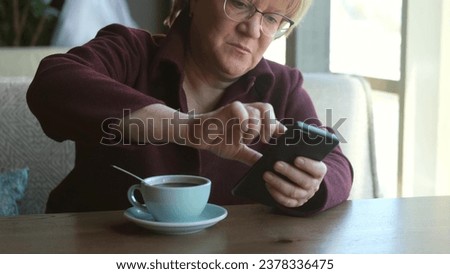 A middle-aged woman with glasses reads the news on a smartphone, makes purchases online using a store app.