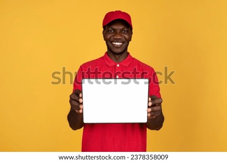 Millennial glad black man courier in red uniform show tablet with empty screen, isolated on orange studio background. Service online, app for delivery, website recommendation Royalty-Free Stock Photo #2378358009