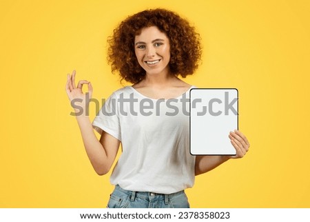 Happy young woman student show tablet with empty screen and ok sign with hand, isolated on yellow background, studio. Good news, recommendation online sale and mobile app, approve website Royalty-Free Stock Photo #2378358023