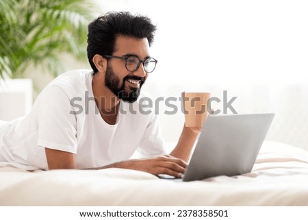Smiling Millennial Indian Guy Relaxing With Laptop And Coffee In Bed, Happy Handsome Eastern Man Browsing Internet On Computer And Enjoying Hot Drink While Resting In Cozy Bedroom, Copy Space