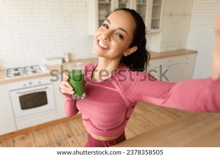 Slim woman in fitwear making selfie posing with smoothie after workout, standing in modern kitchen interior at home. Fitness young lady enjoying green detox drink caring for health and weight loss