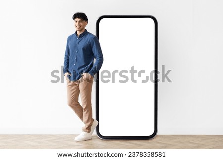 Cheerful middle eastern guy standing near big cellphone with blank white screen, recommending mobile device and new cool app at white studio wall indoor, mockup for website or application design