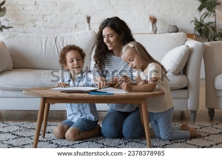 Happy mother helping sweet little daughter and son to draw colorful doodles in paper album, doing school home work at small table, sitting on warm floor, talking, laughing, enjoying daycare activity