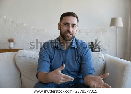 Positive handsome millennial man home head shot. Adult mentor, freelance businessman, blogger speaking at camera, sitting on couch, talking with hand gestures. Video call conversation portrait Royalty-Free Stock Photo #2378379015