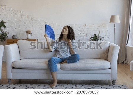 Exhausted frustrated Hispanic woman waving paper handheld fan, cooling hot air, suffering from heat attack, headache, hypoxia, stuffy air, sitting on home couch in living room Royalty-Free Stock Photo #2378379057