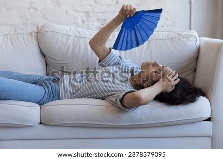 Exhausted sick homeowner woman resting on home sofa, waving handheld paper fan, suffering from fatigue, hypoxia, headache, heating stroke, holding head, swiping sweat Royalty-Free Stock Photo #2378379095