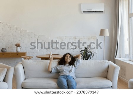 Cheerful relaxed homeowner woman holding AC remote control, starting cooling air conditioning split system at home, resting on sofa with closed eyes, happy smile, enjoying leisure Royalty-Free Stock Photo #2378379149