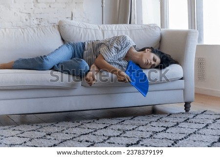 Sick frustrated adult homeowner woman getting heat stroke, lying on sofa, waving handheld paper fan, suffering from hot stuffy air, exhaustion, hypoxia, feeling overheated, bad, ill Royalty-Free Stock Photo #2378379199