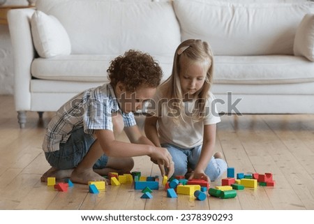 Two focused little kids playing game on warm wooden floor at home, arranging colorful toy building block in living room. Elder brother teaching sister to construct model from heap of colorful pieces Royalty-Free Stock Photo #2378390293