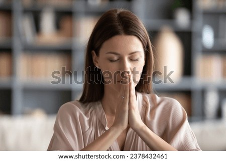 Woman sitting indoors closed eyes folded palms together hands as symbol of request from heart, ask for something personal head shot portrait. Prayer address to God, strong wish, faith, worship concept