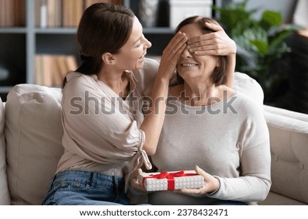 Loving adult grown up daughter prepared for middle-aged mother surprise gift cover her eyes gave present box enjoy moment makes nice for mom on anniversary or birthday, showing love and care concept