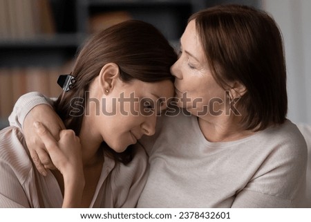 60s mom hugs and kisses on forehead adult daughter seated on couch at home close up. Parent comforting protecting grown up child, showing love provide moral support during heart-to-heart talk concept