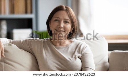 Attractive middle-aged woman sitting on couch smiling looking at camera, old female enjoy photo shooting in modern cozy living room, having calm retired life feels healthy good mood positive emotions