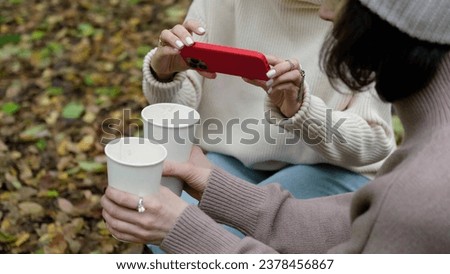 In autumn, two cheerful friends walk through the park during the day, drink coffee and take pictures of glasses of coffee against the background of autumn foliage for social networks