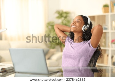 Happy black woman listening music with headphone and laptop relaxing sitting on a chair at home