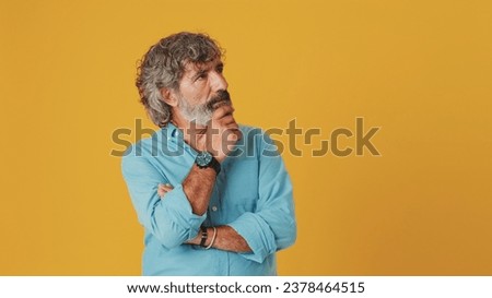Elderly gray-haired bearded man wears a blue shirt, makes a difficult decision, isolated on an orange background in the studio