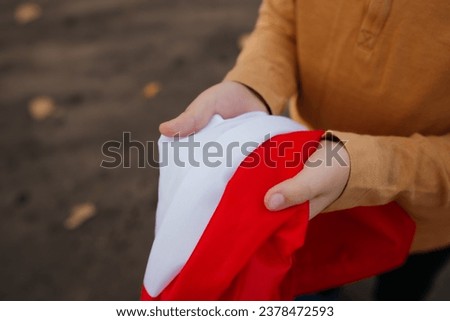 A little boy stands on the road and holds the flag of Poland in his hands. Close-up of child's hands in brown sweater with flag. Concept of national holidays and patriotic events. Independence Day.
