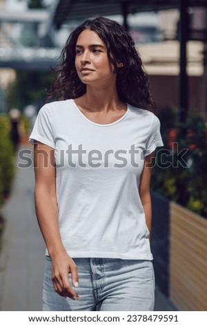 Attractive brunette woman with curly hair in a white t-shirt walks on the street. Mock-up.
