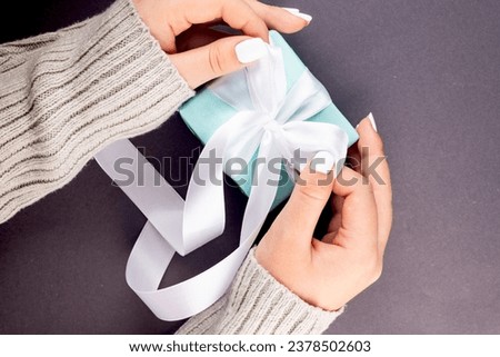 First person top view photo of female hands holding blue gift box with white ribbon on grey background.  My hands cradle a pastel blue gift box