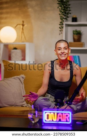 Portrait of a caucasian bald female radio host recording podcast in a home broadcasting studio while sitting on a yellow sofa and looking into the camera. Live stream concept. Copy space.