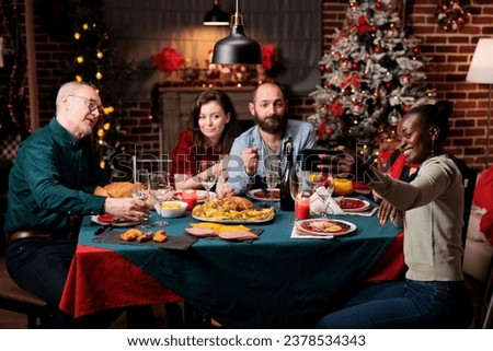 Friends taking pictures on christmas eve, creating beautiful memories of festivity at home. Diverse people having fun with wine and food, enjoying photos for memories at table.