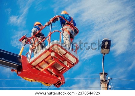 Two electricians from cradle of aerial platform or crane are repairing street lighting lamp. Professional electricians wearing helmets, overalls and insurance work at heights. View of workers from Royalty-Free Stock Photo #2378544929