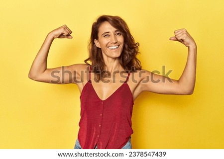 Middle-aged woman on a yellow backdrop showing strength gesture with arms, symbol of feminine power