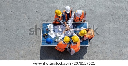 Top view of engineers team, male and female put hands on top over blueprint, solar photovoltaic equipment, road and wind turbine business important infrastructure on solar panel at construction site.