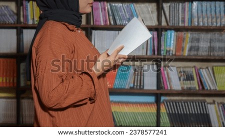 Portrait of Asian hijab woman holding book in front of library bookshelf. Muslim girl reading a book. Concept of literacy and knowledge