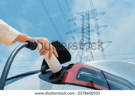 Closeup woman recharge EV electric car battery at charging station connected to electrical power grid tower on sky background as electrical industry for eco friendly vehicle utilization. Expedient Royalty-Free Stock Photo #2378600535