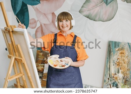 Elderly fun artist woman 50 years old wears casual clothes stand near easel with painting artwork paint listen to music in headphones spend free spare time in living room indoor. Leisure hobby concept
