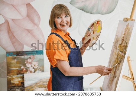 Side view elderly fun happy artist woman 50 years old wearing casual clothes stand near easel with painting artwork paint look camera spend free spare time in living room indoor. Leisure hobby concept