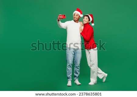 Full body merry young couple man woman wear red casual clothes Santa hat posing doing selfie shot on mobile cell phone isolated on plain green background. Happy New Year 2024 Christmas holiday concept