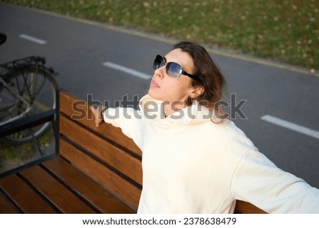 Portrait of a young woman in a modern sun glasses sitting on the wooden bench in a park near a lake enjoying the sunset.Stylish girl in light sports clothes sitting and relaxing on the beach at sunset