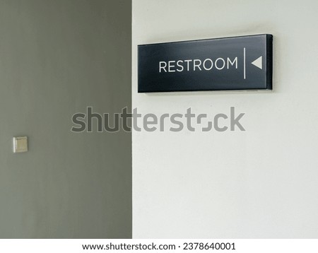 A rectangular black Restroom sign direction with white letters and symbols placed on a white wall of a public area.