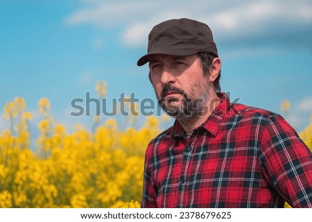 Portrait of confident farmer in blooming canola rapeseed field. Farm worker wearing red shirt and brown trucker's hat at plantation. Selective focus. Royalty-Free Stock Photo #2378679625