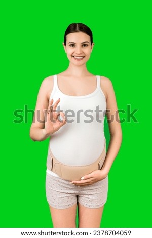Portrait of pregnant woman wearing pregnancy bandage on her belly to make the backache go away and showing okay gesture at green background. Copy space. Orthopedic abdominal support belt concept.