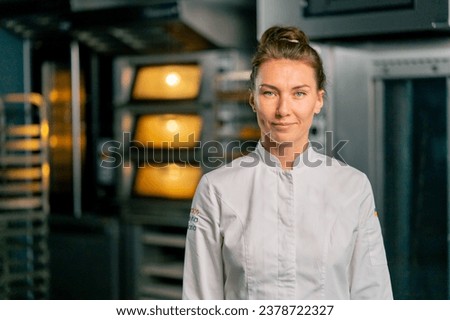 Portrait of a smiling female chef baker in a professional uniform in a bakery against the background of an oven and fresh baked goods