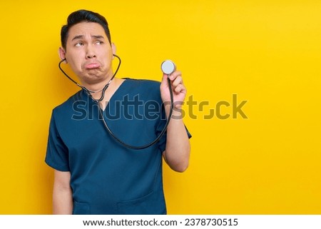 Professional young Asian man doctor or nurse wearing a blue uniform demonstrates using stethoscope isolated on yellow background. Healthcare medicine concept