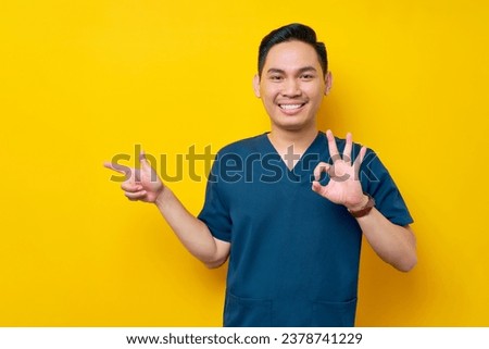 Professional young Asian male doctor or nurse wearing a blue uniform pointing a finger aside and showing an okay sign, looking at camera with smile isolated on yellow background