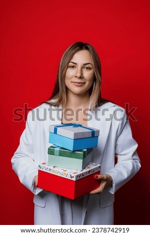 A woman in a stylish white jacket holds boxes of Christmas gifts on a red background. Winter holidays, vacations